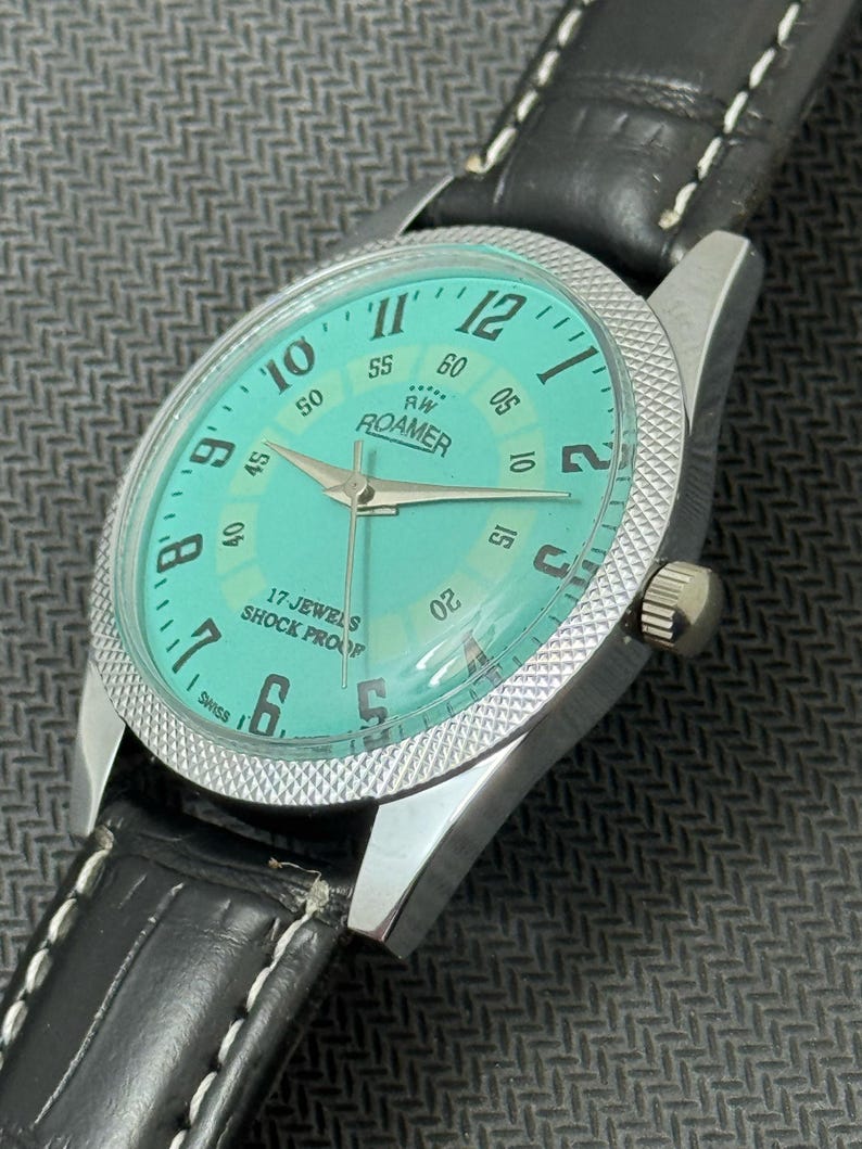 K&ouml;nnte beinhalten: Eine silberne Roamer Armbanduhr mit einem schwarzen Lederarmband und einem t&uuml;rkisblauen Zifferblatt. Das Zifferblatt hat wei&szlig;e Markierungen f&uuml;r die Zahlen und einen wei&szlig;en Sekundenzeiger. Das Zifferblatt der Uhr tr&auml;gt au&szlig;erdem den Text "RW Roamer" und "17 Jewels Shock Proof".