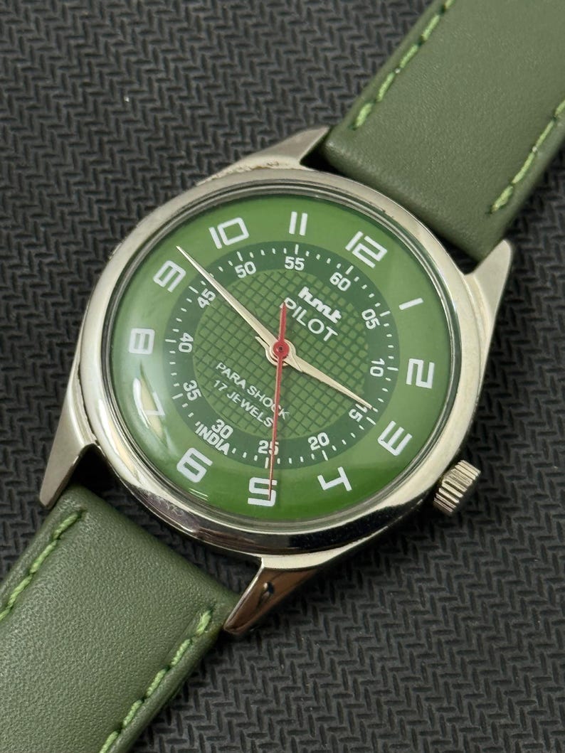Pode incluir: Um rel&oacute;gio de pulso vintage com mostrador verde e caixa prateada. O mostrador exibe algarismos brancos e o texto "hmt PILOT". Possui uma pulseira de couro verde. Os ponteiros do rel&oacute;gio s&atilde;o vermelhos.