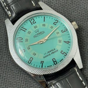 Könnte beinhalten: Eine silberne Roamer Armbanduhr mit einem türkisfarbenen Zifferblatt und einem schwarzen Lederarmband. Das Zifferblatt der Uhr hat weiße Markierungen und den Text "RW Roamer" und "17 Jewels Shock Proof".