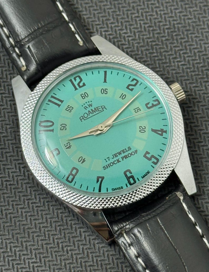 K&ouml;nnte beinhalten: Eine silberne Roamer Armbanduhr mit einem t&uuml;rkisfarbenen Zifferblatt und einem schwarzen Lederarmband. Das Zifferblatt der Uhr hat wei&szlig;e Markierungen und den Text "RW Roamer" und "17 Jewels Shock Proof".
