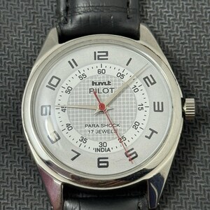 Puede incluir: Un reloj de pulsera plateado con una esfera blanca y una correa de cuero negra. La esfera del reloj tiene marcas negras y una aguja de segundos roja. La esfera del reloj tambi&eacute;n tiene el texto "hmt PILOT PARA SHOCK 17 JEWELS 'INDIA'".