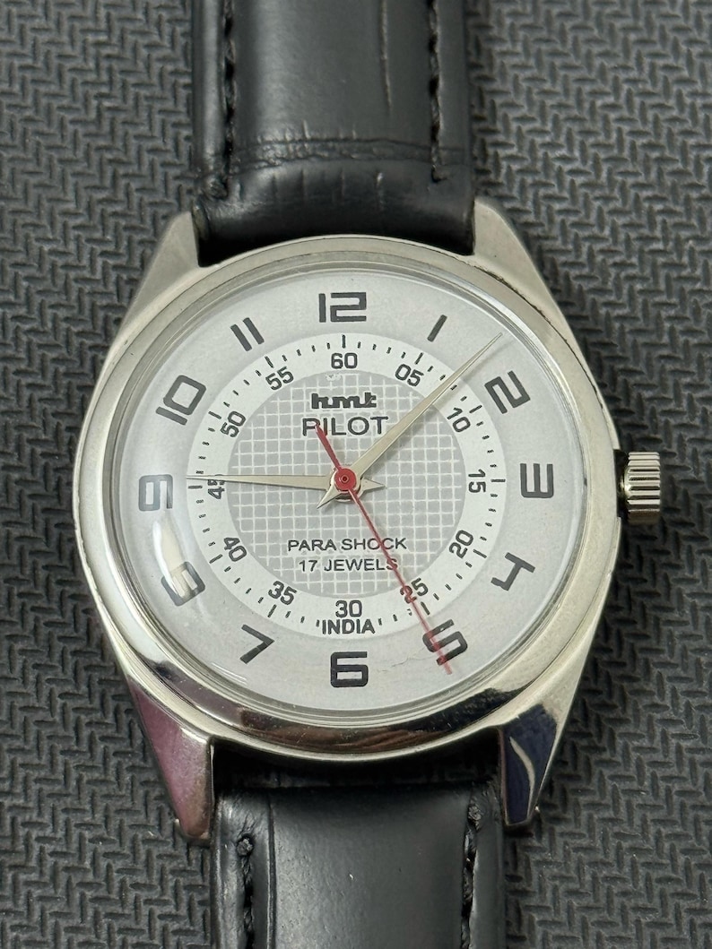 Puede incluir: Un reloj de pulsera plateado con una esfera blanca y una correa de cuero negra. La esfera del reloj tiene marcas negras y una aguja de segundos roja. La esfera del reloj tambi&eacute;n tiene el texto "hmt PILOT PARA SHOCK 17 JEWELS 'INDIA'".
