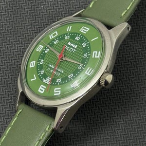 Pode incluir: Um rel&oacute;gio HMT Pilot com mostrador verde, caixa prateada e pulseira de couro verde. O mostrador exibe algarismos brancos e um ponteiro de segundos vermelho. O rel&oacute;gio est&aacute; marcado "PARA SHOCK 17 JEWELS INDIA".