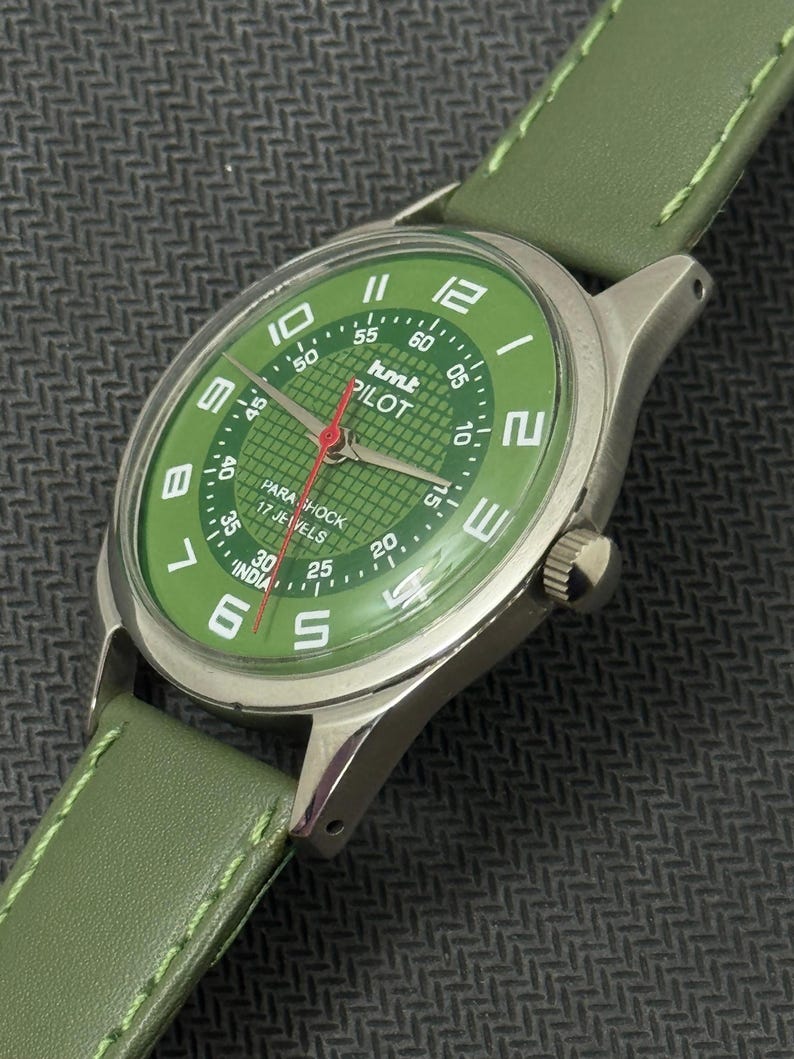 Pode incluir: Um rel&oacute;gio HMT Pilot com mostrador verde, caixa prateada e pulseira de couro verde. O mostrador exibe algarismos brancos e um ponteiro de segundos vermelho. O rel&oacute;gio est&aacute; marcado "PARA SHOCK 17 JEWELS INDIA".