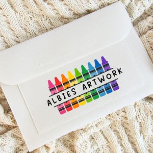 May include: A white envelope featuring a crayon design in rainbow colours. The design includes the text "ALBIES ARTWORK" in black. The envelope is set against a textured, off-white background.