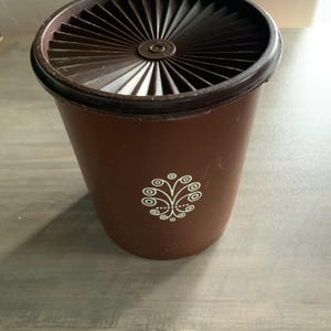 May include: Brown plastic container with a dark brown lid. The lid has a radial design. A white decorative floral design is on the front of the container. The container is cylindrical in shape.