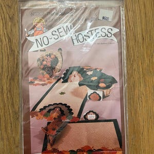 K&ouml;nnte beinhalten: Ein Bastelset zum Thema Thanksgiving in Braun-, Beige- und Gr&uuml;nt&ouml;nen. Das Set enth&auml;lt einen Truthahn, Bl&auml;tter und eine Platzkarte. Der Text auf der Verpackung lautet "No-Sew Hostess" und "6,00 &euro;".