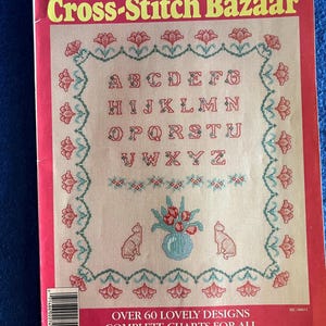 May include: A vintage McCall's Creative Crafts Cross-Stitch Bazaar magazine cover featuring a cross-stitch alphabet design with a vase of tulips and two cats. The magazine cover is red with white text and the title "Cross-Stitch Bazaar". The magazine is volume 27 and costs $3.00.