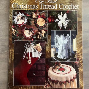 May include: A vintage craft book cover titled "Our Best Christmas Thread Crochet" by Leisure Arts. The cover features various crochet projects, including ornaments, an angel, a table cover, and a Christmas stocking. The projects are displayed with festive decorations.