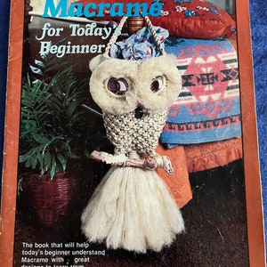 May include: A book titled "Macramé for Today's Beginner" with a picture of a white owl macrame hanging from a wooden dowel. The book cover is orange with white text.