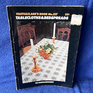 May include: A vintage book titled "COATS & CLARK'S BOOK No. 217 TABLECLOTHS & BEDSPREADS" with a black cover. The cover features an image of a table setting with candles and a floral centerpiece. The book is designed for knitting and crochet.