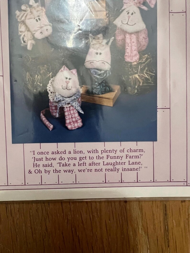 K&ouml;nnte beinhalten: Ein Set aus f&uuml;nf handgefertigten Stofftieren, darunter eine Katze, ein Pferd, ein Schwein und zwei Kaninchen. Die Spielzeuge sind alle aus rosa und wei&szlig;en Stoffen mit gestickten Details. Die Spielzeuge sind auf einer Holzbasis mit einem Zitat &uuml;ber einen lustigen Bauernhof angeordnet.