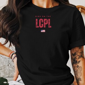 May include: Black t-shirt with the text "DIBS ON THE LCPL" in red letters. The shirt also features a small American flag graphic. The t-shirt is a casual wear item.