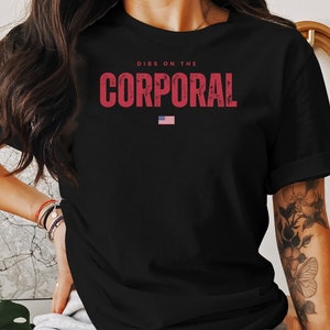 May include: Black t-shirt with the text "DIBS ON THE CORPORAL" in distressed red lettering. A small American flag is printed below the text. The shirt is a crew neck style.