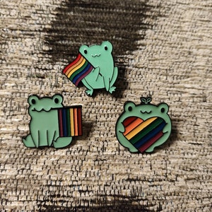 Può includere: Tre spille smaltate con rane verdi con disegni arcobaleno. Una rana tiene una bandiera arcobaleno, un'altra ha un cuore arcobaleno e la terza rana ha una striscia arcobaleno sul suo corpo.