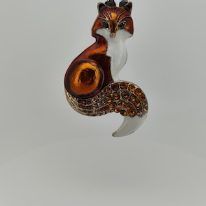 Pode incluir: Uma pequena figura de raposa laranja e branca com uma cauda adornada com joias. A raposa está sentada com a cabeça inclinada para baixo e os olhos fechados. A figura é feita de metal e tem um acabamento brilhante.