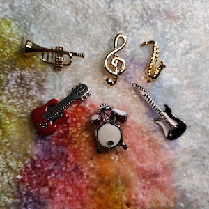 Op de afbeelding: Een set van zes gouden muziekinstrumentencharms, waaronder een trompet, een solsleutel, een saxofoon, een rode elektrische gitaar, een zwarte elektrische gitaar en een drumstel.