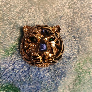 Può includere: Un fermaglio a forma di testa di tigre dorata con occhi di pietre preziose verdi. Il fermaglio è decorato con smalto nero e strass.