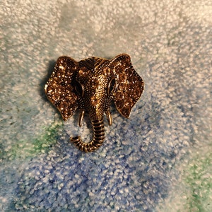 Può includere: Un fermaglio a forma di elefante dorato con strass. La proboscide dell'elefante è arrotolata e le orecchie sono grandi e dettagliate.