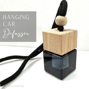 May include: A car air freshener with a black glass cube base, topped with a light wood block and a wooden bead. The black cord is attached to the top. The text "HANGING CAR Difusser" is on a gray background.