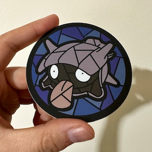 May include: A round sticker with a stained glass design featuring a purple and gray creature with large eyes. The creature is set against a blue and purple background.