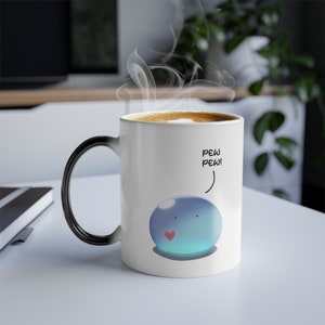 Puede incluir: Una taza de cerámica blanca con asa negra. La taza tiene un personaje de dibujos animados azul y azul claro con un corazón rojo. El texto "PEW PEW!" está escrito encima del personaje.