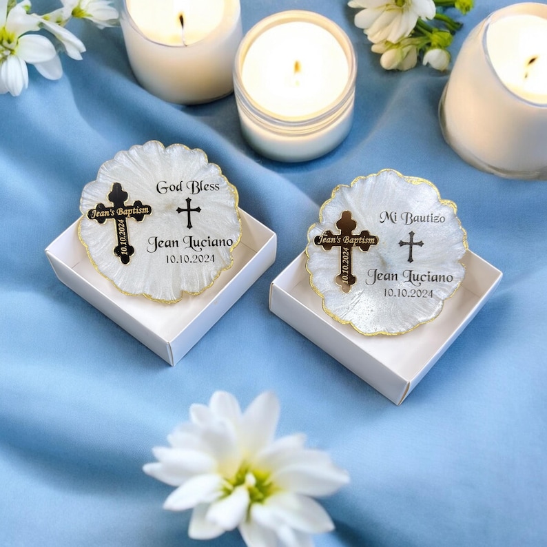 May include: Two white favour boxes with gold trim, each containing a white candle with a gold cross and the text "Jean's Baptism" and "God Bless" or "Mi Bautizo" and "Jean Luciano" with the date "10.10.2024".