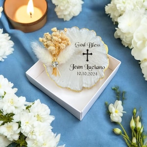 May include: White round resin keepsake with a gold rim, featuring a dried flower bouquet, a cross, and the text "God Bless Jean Luciano 10.10.2024".