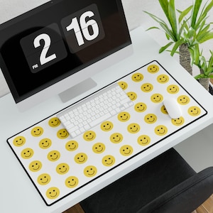 Puede incluir: Un escritorio blanco con un monitor de computadora, un teclado y un ratón. Una alfombrilla de ratón con un patrón de caritas sonrientes amarillas cubre la mayor parte de la superficie del escritorio.