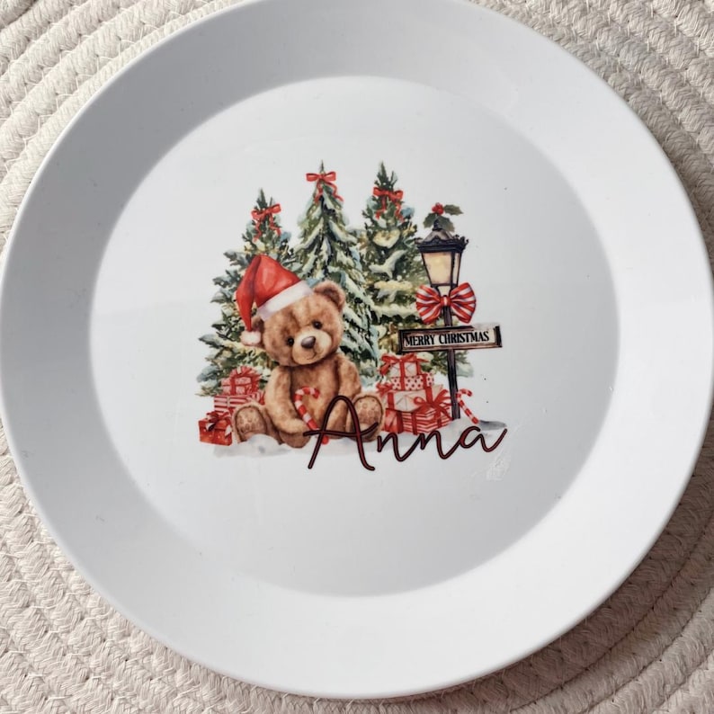 Puede incluir: Un plato de cer&aacute;mica blanca con una ilustraci&oacute;n central de un oso de peluche marr&oacute;n con un gorro de Pap&aacute; Noel rojo, rodeado de &aacute;rboles de Navidad verdes, regalos rojos y una farola con un cartel que dice Merry Christmas y el nombre Anna.