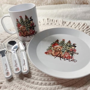 May include: A white Christmas-themed children's dinnerware set including a mug, plate, fork, spoon, and knife. Each item is decorated with a teddy bear in a Santa hat, Christmas trees, and the name "Anna".