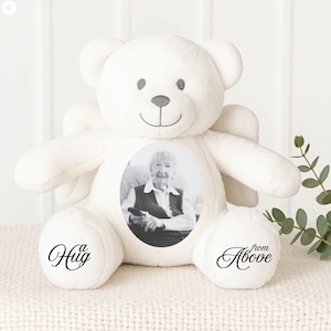 May include: A white plush teddy bear with a photo of a person on its front. The bear has small wings and the words "a Hug" and "from Above" are printed on its paws. The bear is sitting on a white surface.