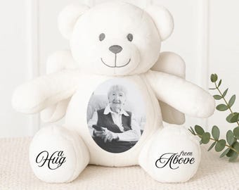 Puede incluir: Un oso de peluche blanco con una foto de una persona en el frente. El oso tiene pequeñas alas y las palabras "a Hug" y "from Above" están impresas en sus patas. El oso está sentado sobre una superficie blanca.