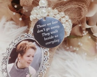 Puede incluir: Un dije de ramo conmemorativo con una foto en un marco plateado adornado. Un dije redondo plateado con la inscripción "Aquellos que amamos no se van, Caminan a nuestro lado todos los días" cuelga sobre la foto. El ramo tiene perlas y arpillera.