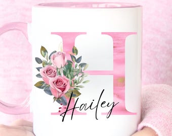 Puede incluir: Taza de cerámica blanca con interior y asa rosas. La taza presenta una gran letra "H" rosa con efecto acuarela, un arreglo floral de rosas rosas y vegetación, y el nombre "Hailey" en escritura negra.