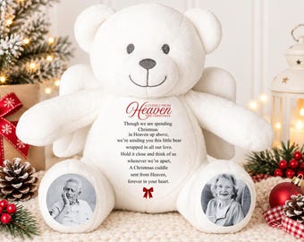 Puede incluir: Un oso de peluche blanco con alas de ángel, que presenta el texto 'A CUDDLE FROM Heaven THIS CHRISTMAS' y un poema sobre la Navidad en el cielo. El oso tiene dos retratos fotográficos circulares en blanco y negro en los pies y un icono de lazo rojo.