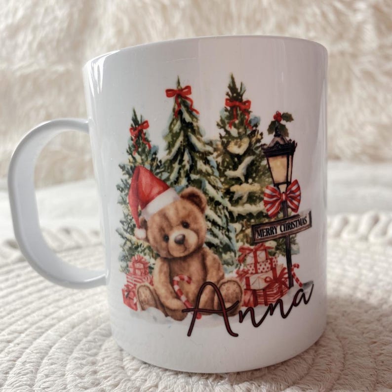Puede incluir: Una taza de cer&aacute;mica blanca con un oso de peluche marr&oacute;n con gorro de Pap&aacute; Noel, rodeado de &aacute;rboles de Navidad, regalos, una farola y un bast&oacute;n de caramelo. Incluye el texto Merry Christmas en un cartel y el nombre Anna escrito debajo.