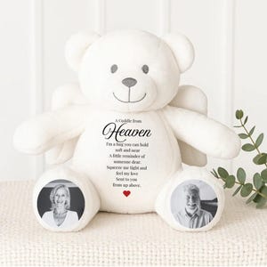 May include: A white teddy bear with angel wings, featuring a smiling face and a heart-shaped nose. The bear has two photo inserts on its feet and text that reads "A Cuddle from Heaven". A touching keepsake.