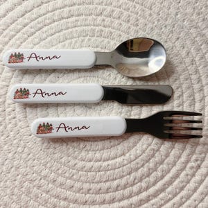 Puede incluir: Un juego de tres cubiertos que consta de una cuchara, un cuchillo y un tenedor. Cada pieza tiene un mango blanco con una peque&ntilde;a ilustraci&oacute;n de un oso y &aacute;rboles, y el nombre Anna impreso en letras cursivas de color rojo oscuro.