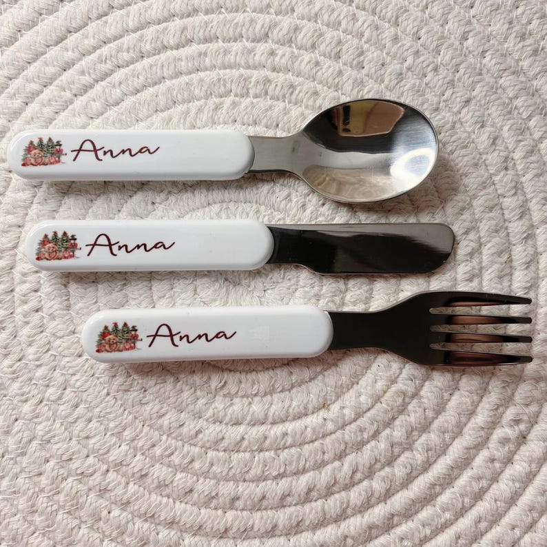 Puede incluir: Un juego de tres cubiertos que consta de una cuchara, un cuchillo y un tenedor. Cada pieza tiene un mango blanco con una peque&ntilde;a ilustraci&oacute;n de un oso y &aacute;rboles, y el nombre Anna impreso en letras cursivas de color rojo oscuro.