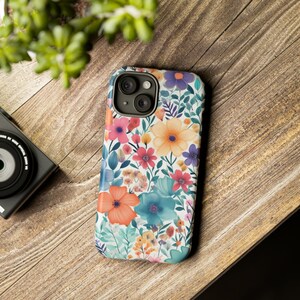May include: A white phone case with a floral pattern featuring pink, orange, yellow, blue, and green flowers. The case has a camera lens cutout and a white background.