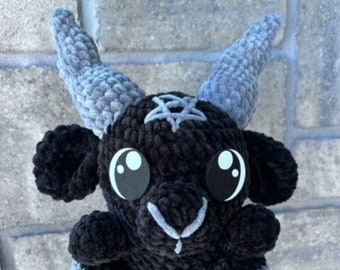 May include: A black crochet plushie of a goat with large, round eyes and gray horns. The goat has a white pentagram on its head and gray accents on its hooves. The plushie is held in a hand against a brick wall.