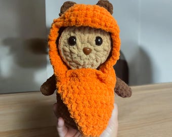May include: A crocheted plush toy in the shape of a character wearing an orange hooded robe. The toy has brown eyes and a brown nose.