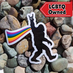 May include: A black silhouette of a Bigfoot with a rainbow coming out of its mouth, giving the rock and roll hand sign. The sticker has the text "LGBTQ Owned" on it.