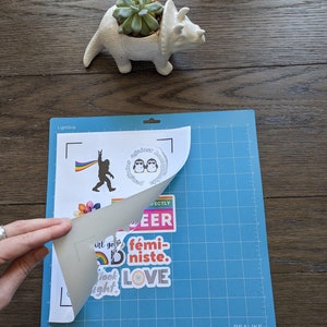May include: A sheet of white paper with several colorful stickers on it. The stickers feature various designs, including a rainbow, a llama, a pair of penguins, and the words "Perfectly Queer", "f&eacute;ministe", and "LOVE". The paper is being peeled off of a blue cutting mat.