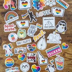 K&ouml;nnte beinhalten: Eine Sammlung bunter Aufkleber mit Regenbogen-Designs und Botschaften der Liebe, Gleichheit und Akzeptanz. Die Aufkleber zeigen S&auml;tze wie "Queer Joy", "Ally", "Pro-body Autonomy", "Good Girl Gone Rad", "Feel", "Love", "Adventure, then Pizza", "Let Me Be Perfectly Queer" und "Squatch the Patriarchy".