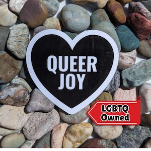 Könnte beinhalten: Ein schwarzer und weißer herzförmiger Aufkleber mit dem Text "QUEER JOY" in weißen Buchstaben. Der Aufkleber befindet sich auf einem Hintergrund aus kleinen, bunten Steinen. Der Aufkleber trägt auch den Text "LGBTQ Owned" in roten Buchstaben.