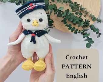 Op de afbeelding: Gehaakt amigurumi speelgoed van een witte vogel die een blauw-witte matrozenhoed draagt met een rode anker. De vogel heeft gele voeten en een rode snavel. Het speelgoed wordt vastgehouden door de handen van een persoon. De afbeelding heeft de tekst "Crochet PATTERN English" op een beige achtergrond.