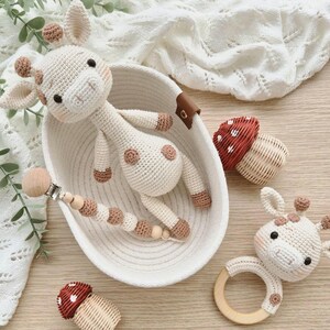 May include: A collection of handmade crochet baby toys, including a giraffe, a pacifier clip, and a rattle. The giraffe is cream-colored with brown spots and accents. The toys are in a white woven basket with a brown leather handle.