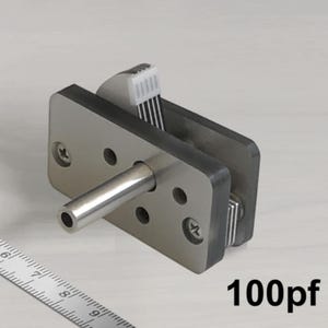 Puede incluir: Un condensador variable de color plateado con una capacidad de 100pF. El condensador tiene una varilla metálica que se extiende desde un lado y una serie de placas metálicas en el otro lado.